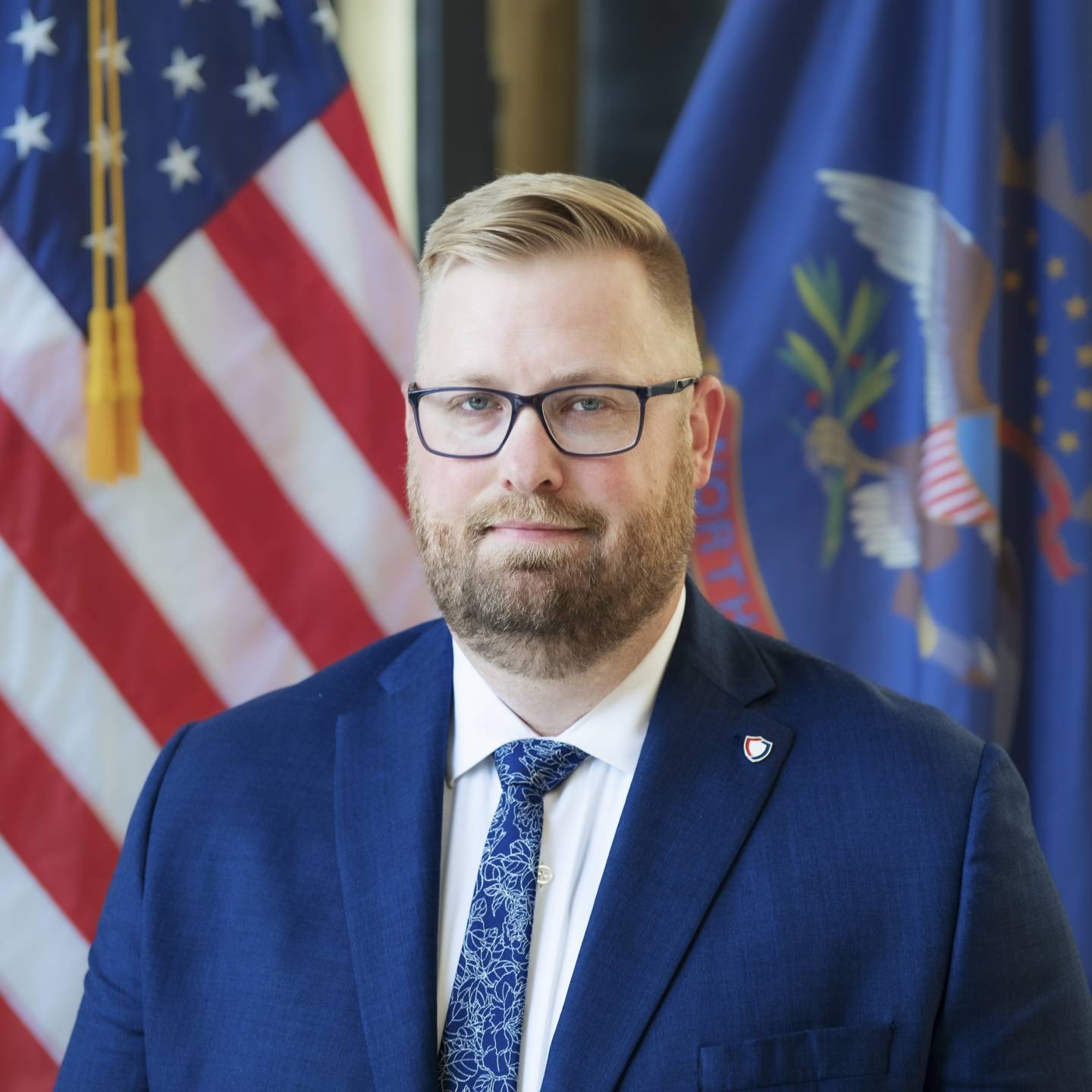 A man in a blue suit and glasses looking forward. Flags in the background.